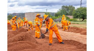Workers in bright orange uniforms and hats shovel sand on a construction site with a rural backdrop. The scene conveys teamwork and diligence.