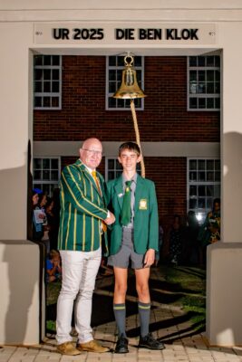 Two people stand under an arch labeled "UR 2025 DIE BEN KLOK," shaking hands. One is older in a striped blazer, the younger in a school uniform. A bell hangs above, with a cheerful atmosphere.
