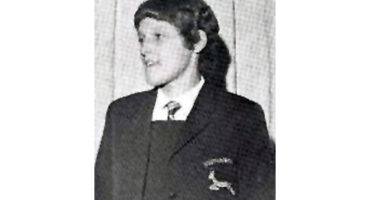 A young person in a Springbok blazer with a badge stands in front of a curtain. They have a short haircut and are smiling slightly, creating a formal and nostalgic tone.