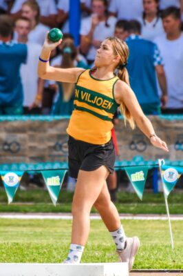 A female athlete in a yellow and green uniform prepares to throw a green shot put at an outdoor event. She stands on a platform, focused, with spectators blurred in the background.