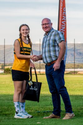 A young girl in a sports uniform shakes hands with a man in casual attire on a grassy field, smiling. She holds a bag labeled "JoJo." An orange banner is in the background.