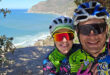A couple wearing bike helmets smiles as they take a selfie together with the ocean in the background.