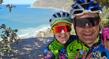 A couple wearing bike helmets smiles as they take a selfie together with the ocean in the background.
