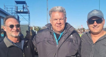 Three men stand together, smiling, at a rugby game, with the field and crowd visible in the background.
