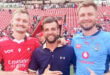 Three men stand smiling at a stadium, two wearing rugby jerseys—one red, one blue. The atmosphere is lively, with a crowd in the background.