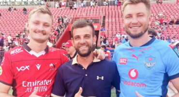 Three men stand smiling at a stadium, two wearing rugby jerseys—one red, one blue. The atmosphere is lively, with a crowd in the background.