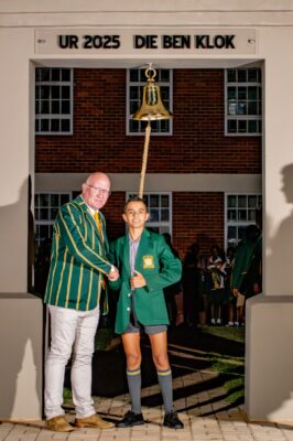 A man and a student in green blazers stand under an arch labeled "UR 2025 DIE BEN KLOK," shaking hands. The student holds a rope tied to a bell.