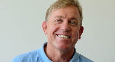 A man wearing a light blue polo shirt sits on a couch in a bright, neutral-toned room.