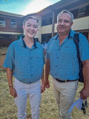 Two people, both smiling, wearing similar blue plaid shirts and light pants, stand in front of a brick building. The setting appears bright and cheerful.