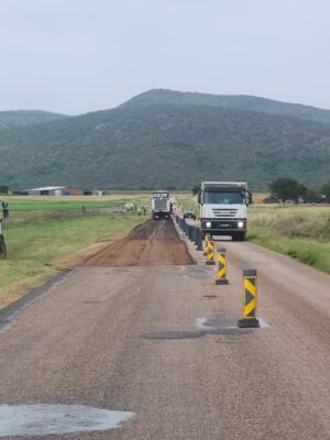 A road construction scene with trucks and safety markers on a rural road, flanked by green fields and a mountain backdrop under an overcast sky.