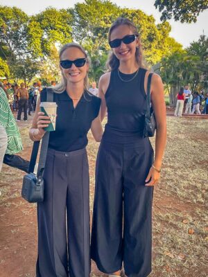 Two women wearing black sunglasses and outfits stand outdoors on grass. One holds a coffee cup, both smiling. Trees and a crowd are visible behind them.