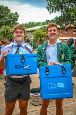 Two smiling students hold blue "BuddyCool" cooler boxes. One wears a tie and shorts, the other a green blazer. Background shows a lively schoolyard.