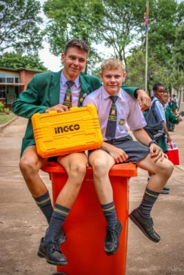 Two male students in uniforms sit on an orange trash bin, smiling with a bright yellow tool case labeled "ingco." Background shows trees and buildings.