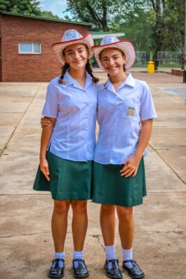 Two smiling girls in white shirts and green skirts stand arm-in-arm, wearing straw hats, on a school courtyard with trees and a brick building.