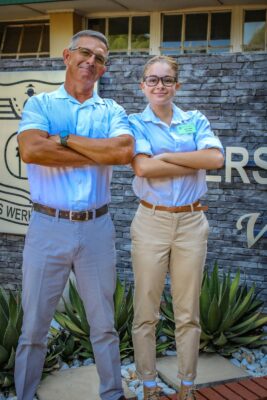 A man and a young woman stand confidently with arms crossed, smiling. They wear light blue shirts and beige pants, in front of a stone wall with text.