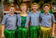 Four young men in school uniforms and festive hats, smiling and standing close. They wear shiny green skirts, exuding a cheerful, playful vibe outdoors.