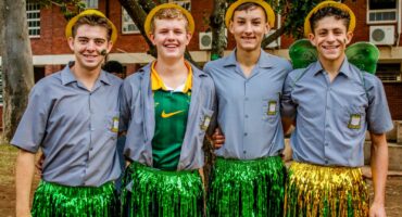 Four young men in school uniforms and festive hats, smiling and standing close. They wear shiny green skirts, exuding a cheerful, playful vibe outdoors.