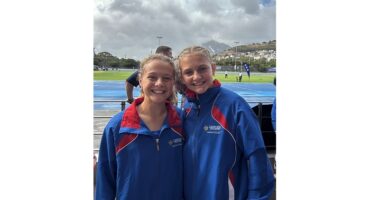 Two young athletes in blue and red tracksuits smile at the camera, standing on an outdoor track, with hills and a cloudy sky in the background.