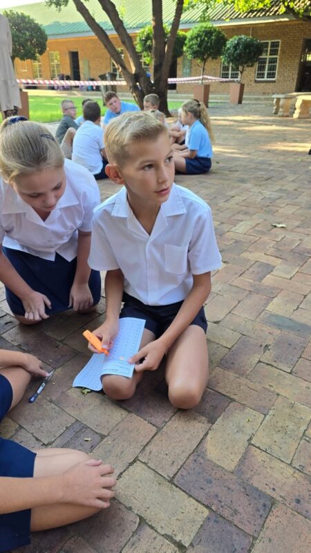 A boy in a school uniform sits on brick pavement holding paper and a highlighter, with peers around him, engaged in outdoor learning activities.
