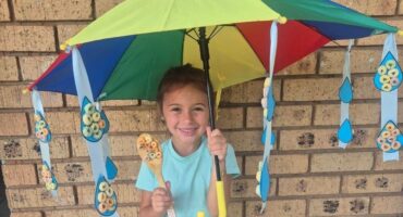 A smiling child holds a colorful umbrella with cereal-decorated ribbons. She stands in front of a brick wall, holding a spoon. The scene is playful and cheerful.