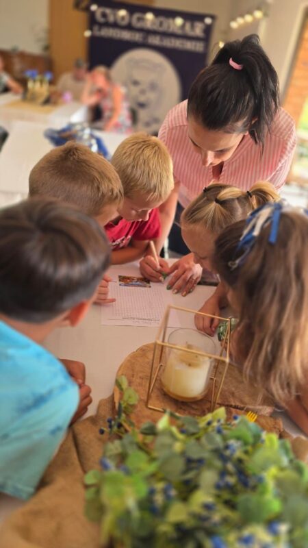 A group of young children and a woman are gathered around a table, focused on a worksheet. A flower arrangement and candle holder add a cozy touch.