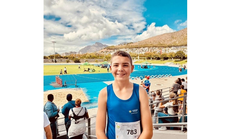 Smiling boy in a blue athletic tank top stands in a stadium with a bright blue track, mountains in the background, and a crowd watching a sporting event.
