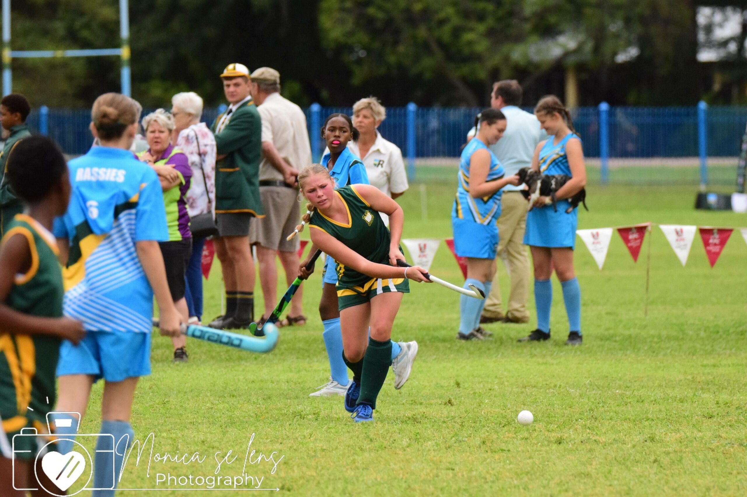 A field hockey player in a green and yellow uniform intensely focuses on hitting the ball. Teammates and opponents in blue jerseys are visible, with spectators in the background. The scene conveys competition and teamwork.