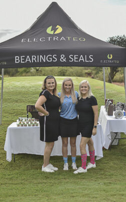 Three women stand smiling under a tent labeled "Electrateq Bearings & Seals" on a grassy field. They are wearing sports attire and colorful socks.