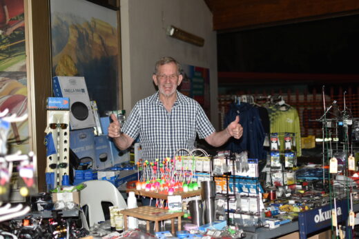 A man in a checked shirt stands behind a fishing gear display, giving two thumbs up. The table is filled with colorful floats and various fishing supplies.