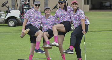 Four women on a golf course pose playfully with raised legs. They wear matching pink patterned shirts, bright pink socks, and shorts, conveying a fun and cheerful vibe. Golf carts and clubs are visible in the background.