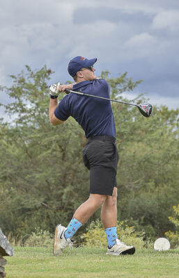 A golfer in mid-swing stands on a lush course, wearing a navy shirt, black shorts, and blue socks. The sky is overcast, suggesting a calm yet focused mood.