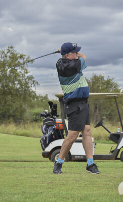 A golfer swings a club on a lush green course, wearing a striped polo and blue socks. A golf cart and cloudy sky are in the background, conveying focus and action.