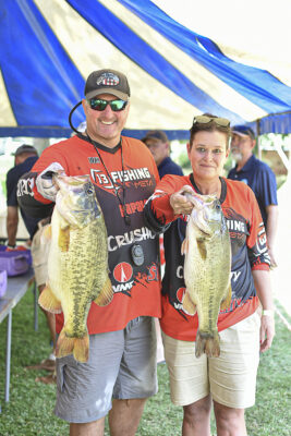 Two people stand under a blue and white tent, smiling and holding large bass fish. They wear matching red fishing shirts, conveying excitement.