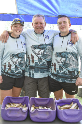 Three people wearing matching fishing shirts with a large fish graphic stand smiling under a blue tent. Purple bins in front hold various small fish.