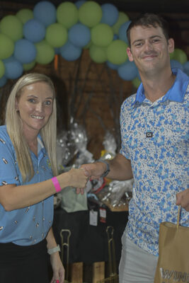 A woman and a man smiling while shaking hands at an event. The background features a decorative arch of blue and green balloons, with gift bags visible.