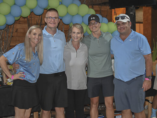 Five people smiling, standing closely together indoors. They are casually dressed in short-sleeved shirts and shorts. Background features blue and green balloons.