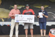 Three men stand holding a large check for R5,000 at a fishing competition. They appear pleased, with a fish-themed banner in the background.