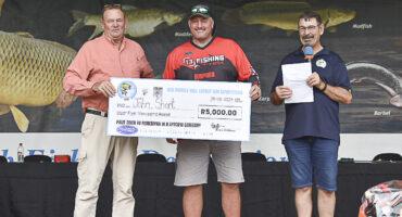 Three men stand holding a large check for R5,000 at a fishing competition. They appear pleased, with a fish-themed banner in the background.