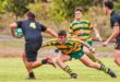 Rugby match action with a player in yellow and green tackling an opponent in black holding the ball. Two other players watch, showing intensity and focus.