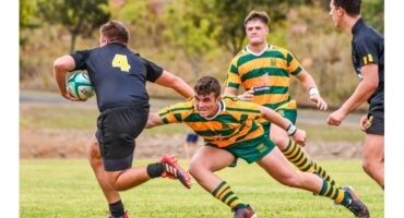 Rugby match action with a player in yellow and green tackling an opponent in black holding the ball. Two other players watch, showing intensity and focus.