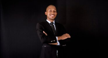 A confident man in a black suit and tie stands against a dark background, smiling with arms crossed. The tone is professional and approachable.