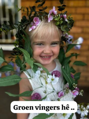 A joyful child with blonde hair smiles warmly, adorned with a crown of green leaves and purple flowers. The text reads, "Groen vingers hê ..".