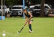Young female field hockey player in green and yellow uniform focuses on ball control in a grassy field. Parked cars and trees are in the background.