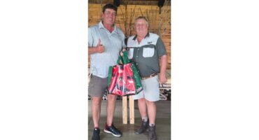 Two men smiling indoors, one giving a thumbs up, holding colorful gift bags. They stand on a wooden floor with a rustic background. Casual, cheerful mood.