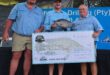 Three men wearing blue shirts stand together outdoors holding a large check for fifty thousand rand. The middle man holds a fish, smiling. It's an award ceremony.