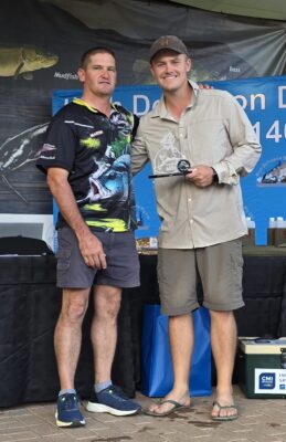 Two men pose happily at a fishing event. The man on the right holds a trophy. A banner with fish images is displayed in the background.