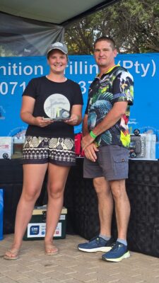A smiling young woman holds a trophy next to a man, both in casual attire. A blue banner in the background suggests a competition setting.