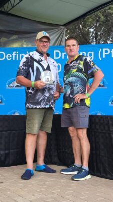 Two men stand on a stage in front of a blue backdrop with fish graphics. Both wear fish-themed shirts, looking relaxed and content.