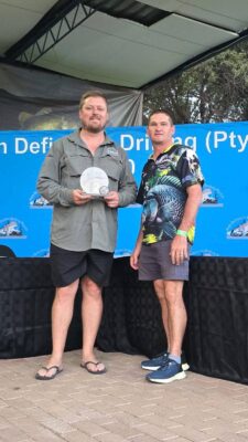 Two men pose with an award under a tent. One holds a trophy, wearing casual clothes and sandals. The backdrop is blue and reads "Drilling."