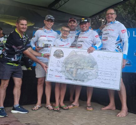 A group of men and a child smiles while holding a large check for 10,000 Rand, celebrating a fishing tournament win under a canopy. They wear branded fishing shirts, conveying excitement and achievement.